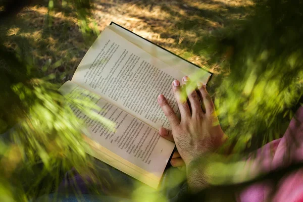 Kot pantolonlu ve pembe gömlekli beyaz bir adam ormanda bir ağacın yanında oturmuş siyah bir kitap okuyor.