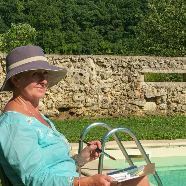 A lady artist sits in sunshine beside a swimming pool working on the painting she started earlier in the day.