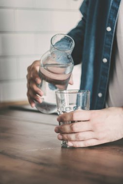 Adam kendine bir bardak temiz su dolduruyor. Mutfak ahşap masasında bir sürahi su. Bir şişe su. Sağlıklı yaşam tarzı kavramı