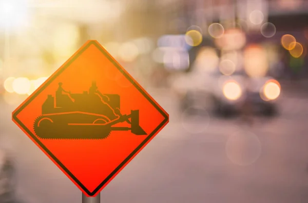 Construction tractor warning sign on blur traffic road with colorful bokeh light abstract background. — Stock Photo, Image