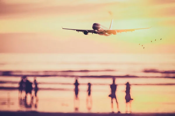 Airplane flying over blur people relax on tropical sunset beach with bokeh sun light wave abstract background. — Stock Photo, Image