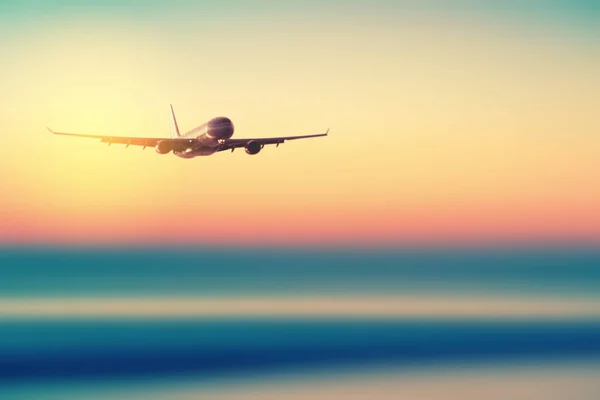 Airplane flying over blur tropical beach with smooth wave and sunset sky abstract background. — Stock Photo, Image