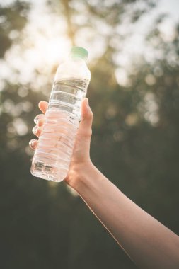 Kadın, elinde bir şişe su ile doğada yeşil bulanık bir yaprak ve gün batımı ışığı soyut bir arka plan..