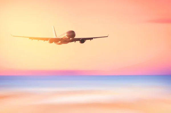 Airplane flying over blur tropical beach with smooth wave and sunset sky abstract background. — Stock Photo, Image