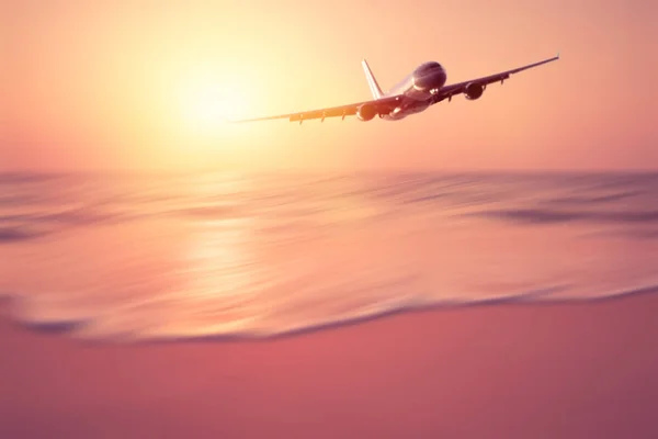 Airplane flying over blur tropical beach with smooth wave and sunset sky abstract background. — Stock Photo, Image