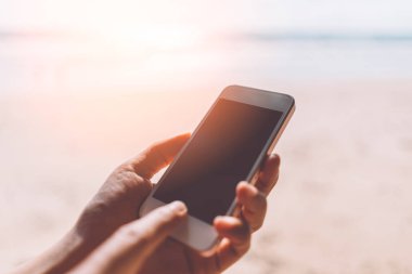 Tropikal günbatımı plaj soyut arka plan üzerinde akıllı telefon kullanarak kadın eli kapatın. Teknoloji iş ve seyahat tatil konsepti. 