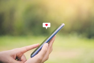 Açık hava parkı arka planında kalp ikonu olan akıllı bir telefon kullanarak kadının elini tutun. Teknoloji ve seyahat tatil konseptinin kopyalama alanı. Klasik ton filtre efekti renk biçimi.