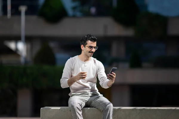 young company worker next to skyscraper buildings reading messages on mobile phone and drinking coffee on break from work