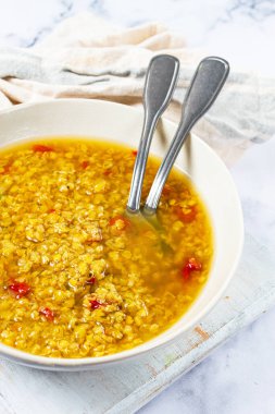 Kırmızı biber taneleriyle süslenmiş sıcak, altın mercimek çorbasıyla dolu bir kase, iki metal kaşıkla birlikte, ahşap bir yüzeyde oturur. Çorba davetkar ve lezzetli görünüyor..