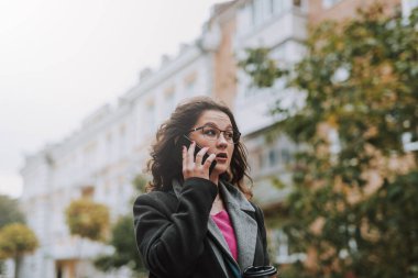 Düşünceli, güzel, gözlüklü bir kadın elinde bir fincan kahveyle sokakta yürürken telefonla konuşuyor.