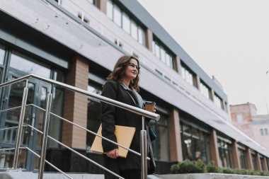 Büyük bir kitap ve bir fincan kahveyle modern binanın yanında duran ve gülümseyen oldukça mutlu genç bir bayan.