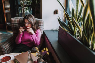 Güzel bayan kafede tek başına oturuyor ve elinde sıcak içkinin tadını çıkarırken başka tarafa bakıyor.