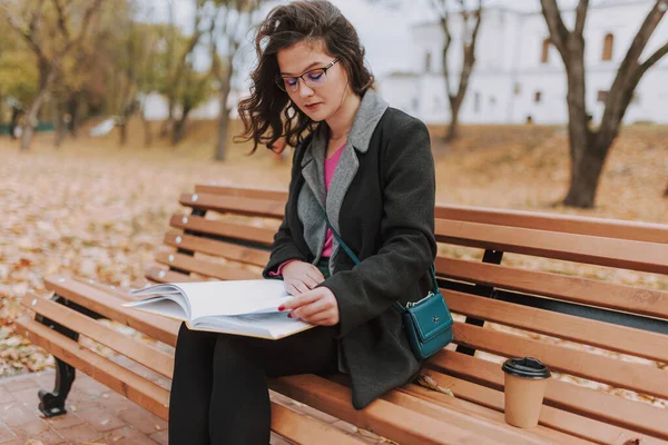 坐在外面的长椅上 穿着外套和眼镜看书的迷人的年轻女士 — 图库照片
