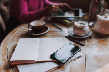 Akıllı telefon ve bir fincan çay masanın üstünde, arka planda bir kadın çalışıyor.
