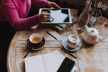 Tahta masadaki çay fincanının en üst görüntüsü ve kahve evinde dijital tablet kullanan kadın.