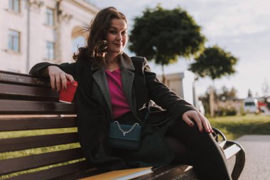 Kentteki sıcak içkinin tadını çıkarırken bankta oturan çantalı mutlu genç bayan.