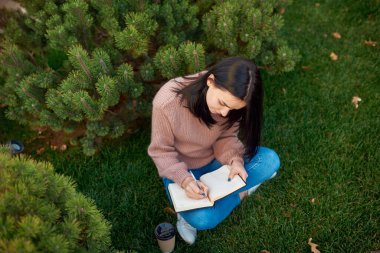 Kalın bir defter ile kayıt yapan genç bir kadının yeşil çimlerin üzerinde otururken kozalaklı ağaçlarla çevrili görüntüsü.