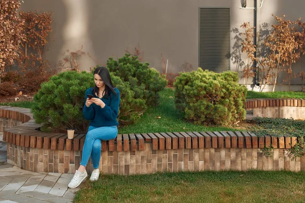 Düşünceli ve ciddi bir genç kadın cep telefonuyla vakit geçiriyor ve dışarıda oturuyor.