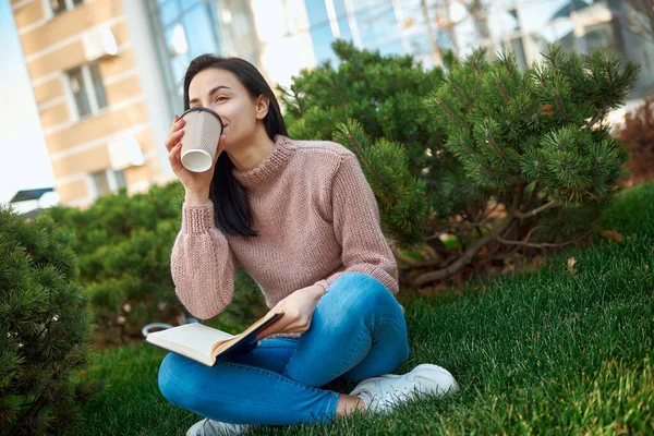 Pretty young woman with a thick daybook on her knees enjoying hot drink on a green lawn near tall modern building