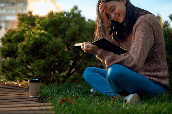 Young beautiful female sitting on a sward near bushy dwarf pine and leaning over a book with smile