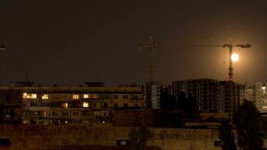 Arka planda Ay 'ın hareketi ön planda çeşitli vinçler ve konut binaları olan büyük bir inşaat sahası. Geceyarısı çekim.