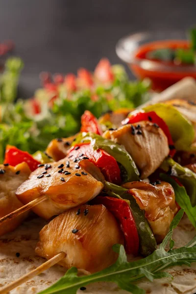 Serve food in a restaurant. Food poster - large portion of chicken kebab with black sesame seeds and ketchup. Lettuce, red bell pepper, cucumber and tomatoes. Served on a black stone serving board.