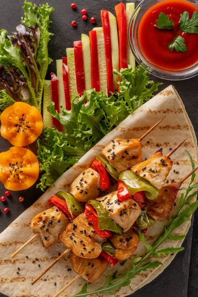 Serve food in a restaurant. Food poster - large portion of chicken kebab with black sesame seeds and ketchup. Lettuce, red bell pepper, cucumber and tomatoes. Served on a black stone serving board.