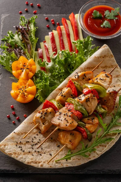 Serve food in a restaurant. Food poster - large portion of chicken kebab with black sesame seeds and ketchup. Lettuce, red bell pepper, cucumber and tomatoes. Served on a black stone serving board.