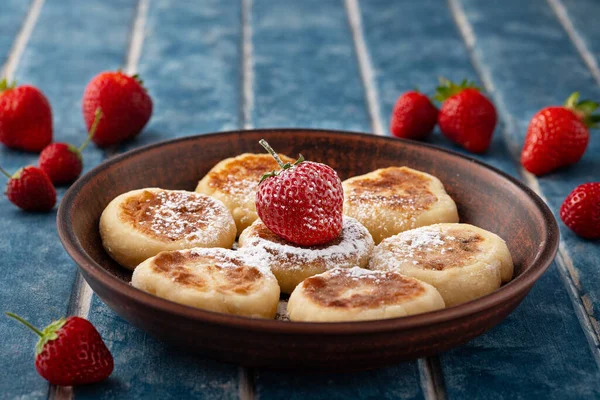 Cottage cheese pancakes, pancakes, cheesecakes with strawberries in a clay plate on a background of blue wooden boards. Ukrainian and Russian cuisine. Homemade food. Tasty breakfast.
