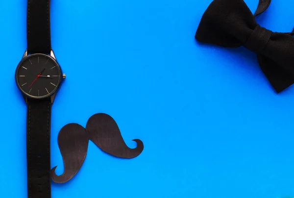 International Fathers Day. Mens holiday. Greeting card for dad. Wristwatch, paper mustache and bow tie on a blue background. Mens Accessories. Place for text. Flat lay. Top view.