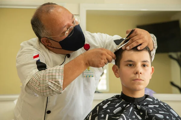 Genç çocuğun saçını kesen, salgın yüzünden koruyucu maske takan mutlu Latin berber.