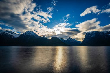Bulutlu akşam gökyüzü, sakin göl suyu yüzeyi ve dağlar. 