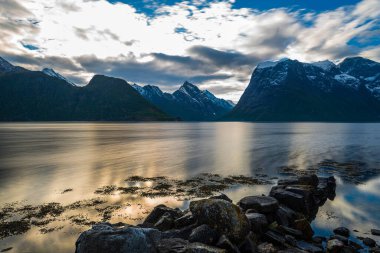 Bulutlu akşam gökyüzü, sakin göl suyu yüzeyi ve dağlar. 