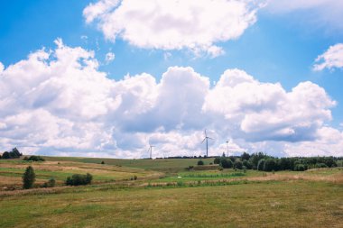 Bulutlu bir gökyüzü ve üzerinde rüzgar jeneratörlerinin durduğu tepelik bir alan.