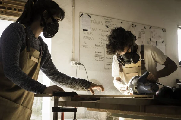 Young couple working together as carpenter in a small carpentry workshop. Young man slicing a piece of timber with a wood saw machine wearing a face mask for the dust. Couple working together