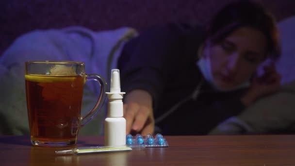Zieke vrouw in medisch masker met moeite pakt pillen van tafel met thermometer — Stockvideo