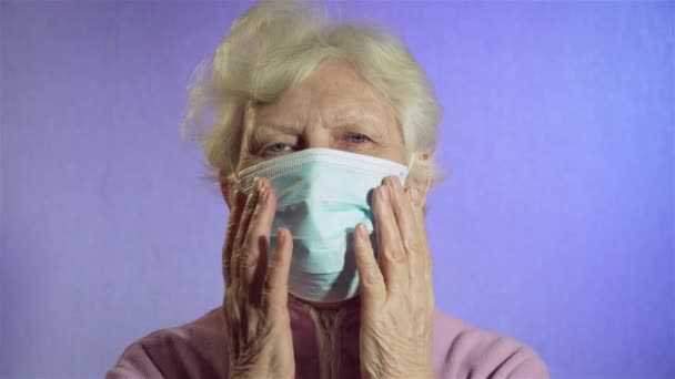 Die ältere Frau berührt ihr Gesicht mit einer medizinischen Schutzmaske und blickt in die Kamera. Coronavirus in Quarantäne für alte Menschen — Stockvideo