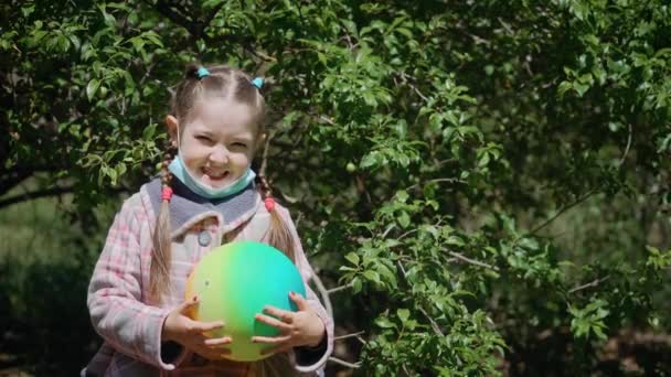 Schattig klein meisje met naar beneden gericht beschermend masker met bal in handen glimlachend naar de camera — Stockvideo