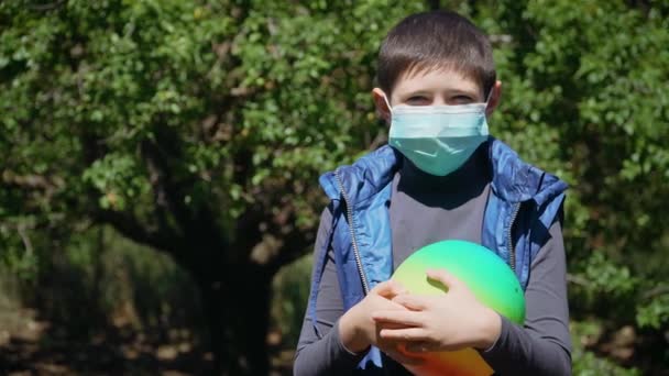 Niño aburrido en máscara protectora en la cara en casa en el patio trasero en cuarentena con bola de colores — Vídeo de stock