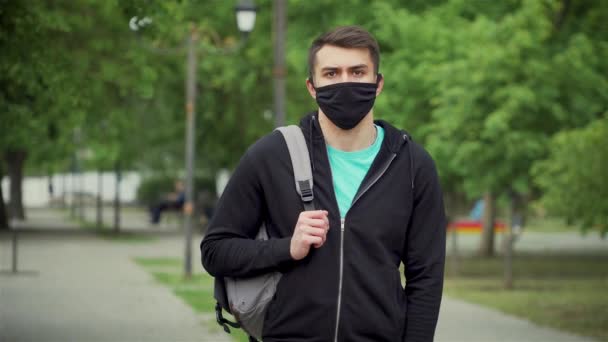 Junger Mann mit Umhängetasche steht zur Coronavirus-Prävention mit Mundschutz im Stadtpark — Stockvideo