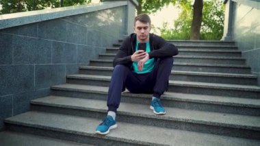 Şehir parkında merdivenlerde oturan genç adam kablosuz bağlantı kullanarak cep telefonundan mesajlar yolluyor.