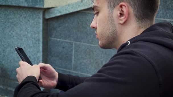 Ung kaukasisk amerikansk man som håller smartphone enhet sms meddelande sitter vid trappor i stadsparken — Stockvideo