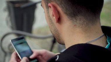 Genç ve çekici bir adamın sosyal ağları parktaki bankta cep telefonuyla izlemesini kapatın.