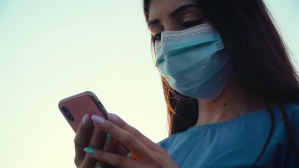 Retrato de mulher em máscara médica protetora digitando mensagens no telefone celular enquanto a pandemia do vírus corona começou e seu longo cabelo escuro está soprando no vento — Vídeo de Stock