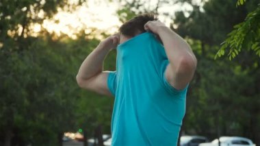 Yavaş çekim çekici genç adam modeli görünümü tişörtünü çıkarır ve atletik gövdesini gösterir ve gün batımında kendine güvenen bir şekilde kameraya bakar.
