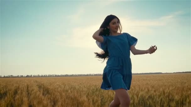 Schöne junge Frau mit langen dunklen Haaren in blauem Kleid läuft im Weizen- oder Gerstenfeld bei Sonnenuntergang in Zeitlupe — Stockvideo