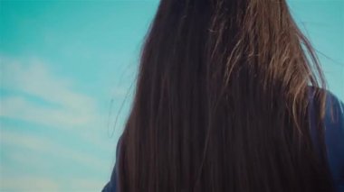 Siyah saçlı, gülümseyen genç bir kadın kameraya dönüp mavi gökyüzünün altında yürüyor.