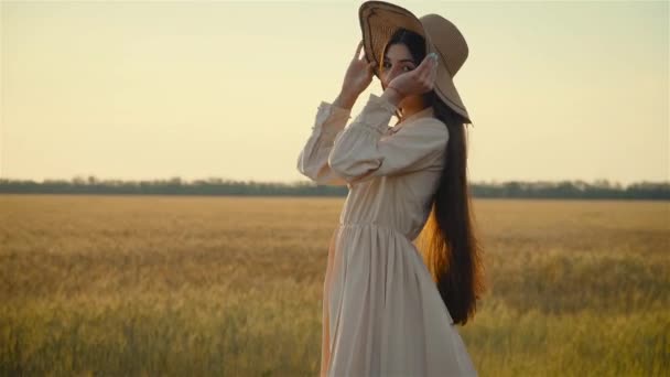 Mujer europea joven y sonriente confiada en vestido ligero se pone el sombrero y sonríe. Disfrutando de la naturaleza al atardecer cerca del campo agrícola — Vídeo de stock