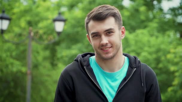 Retrato de un joven adulto sonriente mirando a la cámara y de pie en el parque — Vídeos de Stock