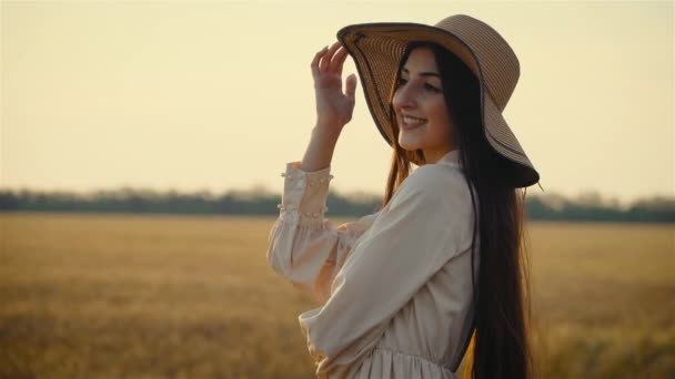 Hermosa mujer sonríe parada sola en sombrero de paja parada sola al atardecer cerca de campo rural agronómico con cultivos — Vídeo de stock